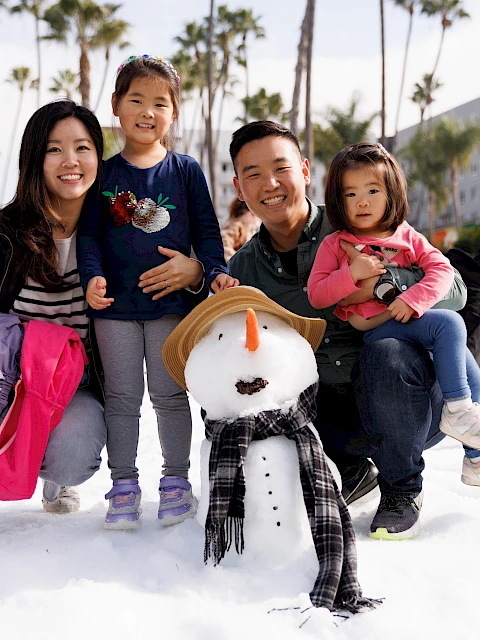 A family poses with a snowman in a snowy park area, surrounded by palm trees and other people enjoying the snow.