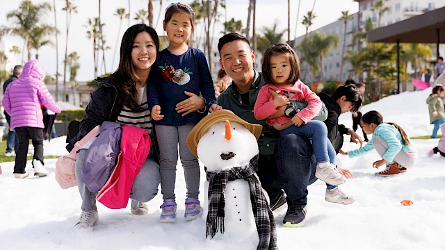 A family poses with a snowman in a snowy park area, surrounded by palm trees and other people enjoying the snow.