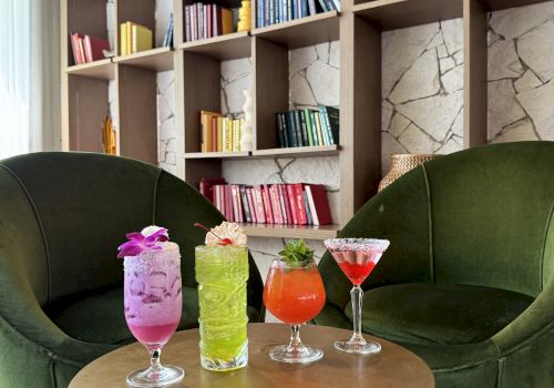 Lounge area with two green chairs, colorful drinks on a table, and a bookshelf with books organized by color in the background.