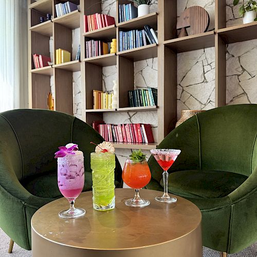 Lounge area with two green chairs, colorful drinks on a table, and a bookshelf with books organized by color in the background.