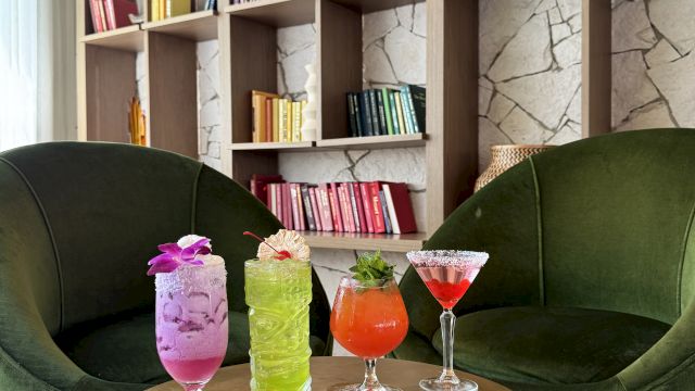 Lounge area with two green chairs, colorful drinks on a table, and a bookshelf with books organized by color in the background.