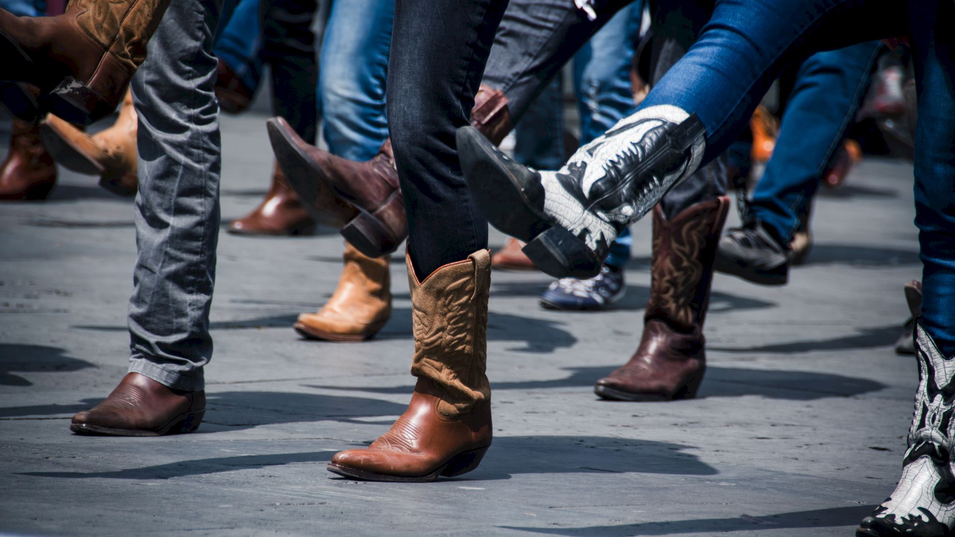 People are line dancing, wearing various styles of cowboy boots, with legs lifted in motion on a concrete surface.