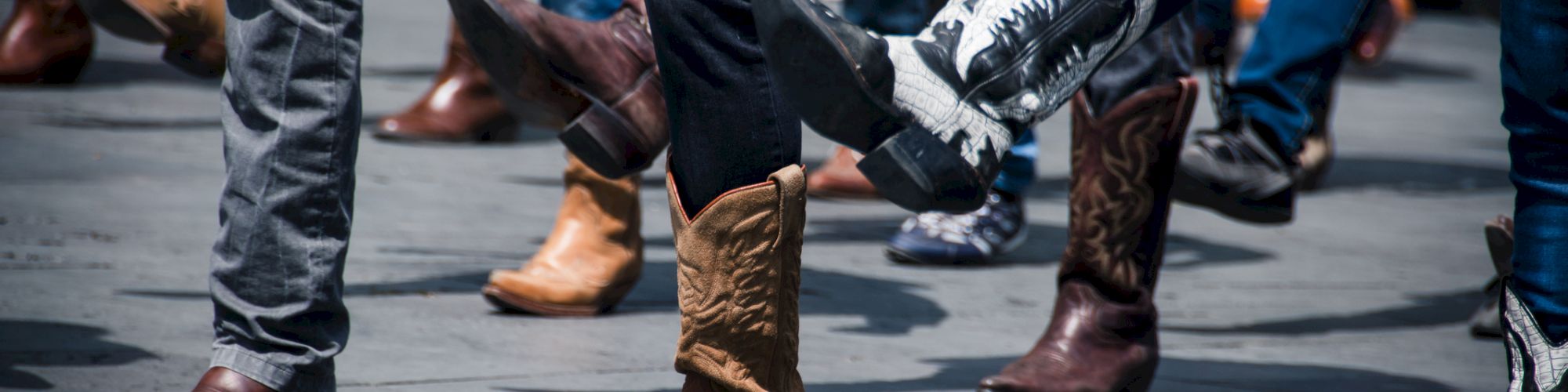 People are line dancing, wearing various styles of cowboy boots, with legs lifted in motion on a concrete surface.