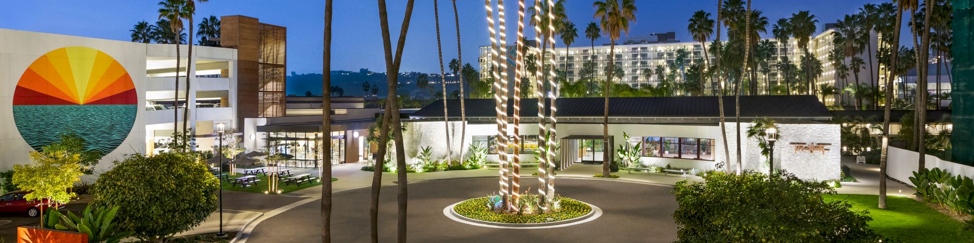 The image depicts an outdoor scene of a modern building complex, surrounded by tall palm trees, decorative lighting, and landscaped gardens at dusk.