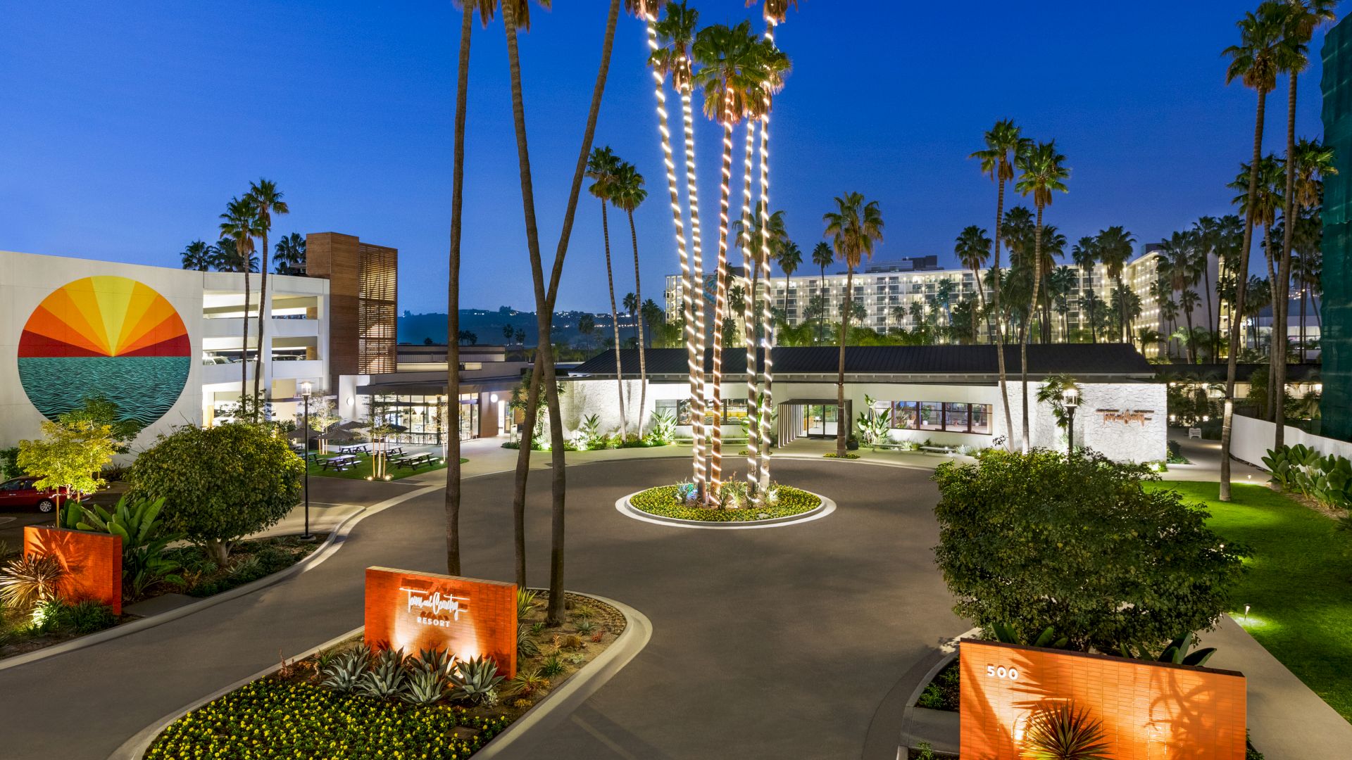The image depicts an outdoor scene of a modern building complex, surrounded by tall palm trees, decorative lighting, and landscaped gardens at dusk.