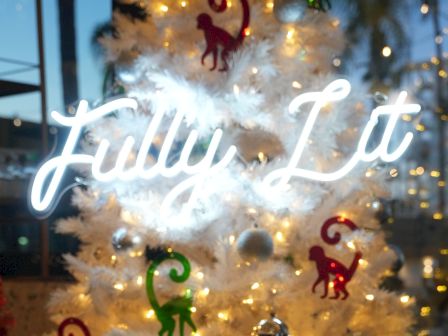 A white Christmas tree adorned with lights and colorful decorations, plus a neon sign reading &ldquo;Fully Lit&rdquo; in cursive across the glass.