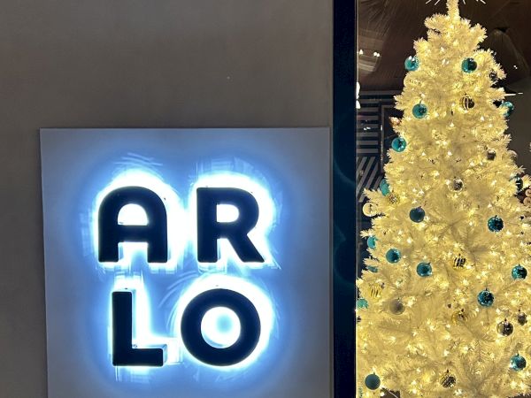 The image shows a sign with illuminated "ARLO" letters next to a white Christmas tree adorned with blue ornaments and white lights.