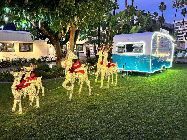 Lit-up reindeer decorations with red ribbons stand on grass beside a blue and white holiday trailer; trees and buildings are in the background.