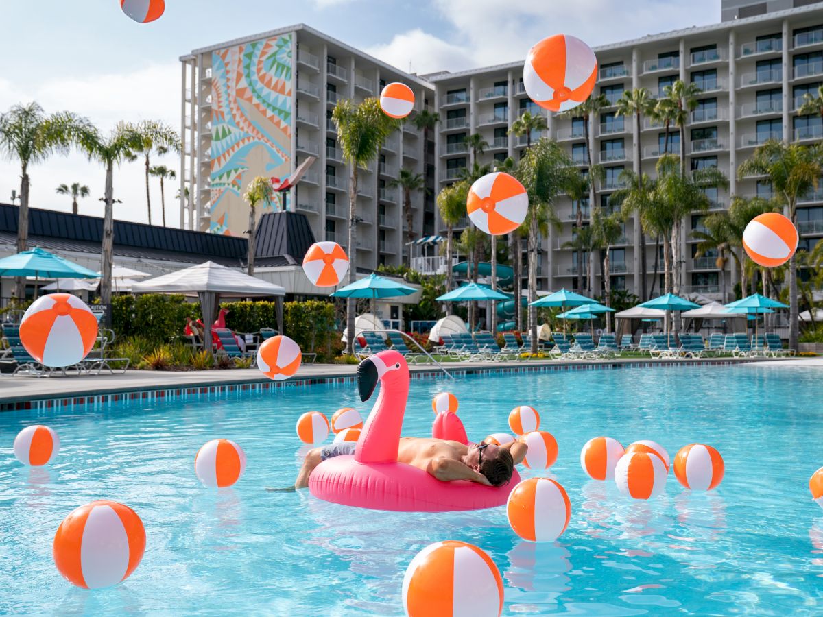 People lounging and floating in a hotel pool filled with orange-and-white beach balls; a pink flamingo float and palm trees backdrop.