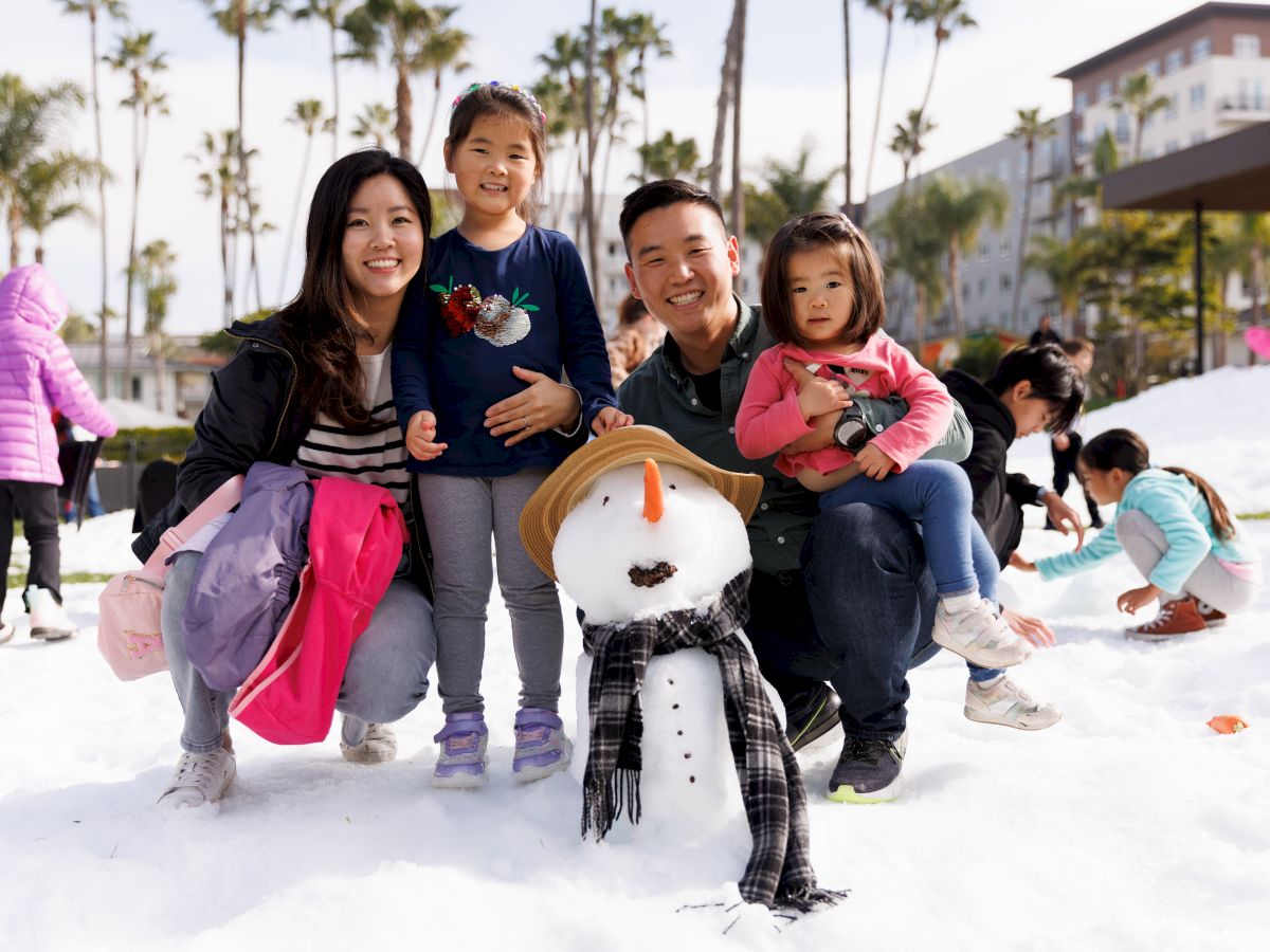 A smiling family poses with a small snowman on a sunny beachy-white snowy yard; kids play nearby, palm trees and buildings in background.