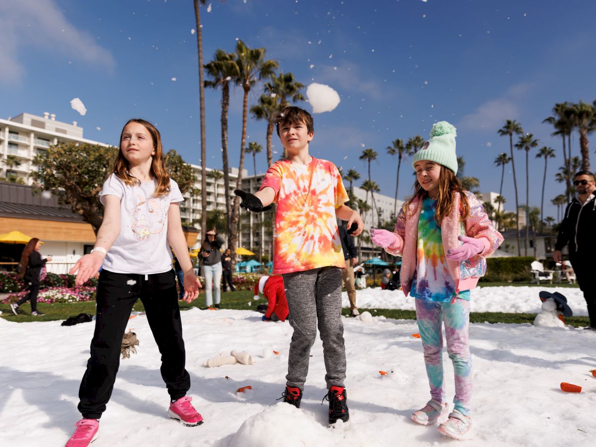 The image shows three children playing in the snow at a park, with palm trees and buildings in the background.
