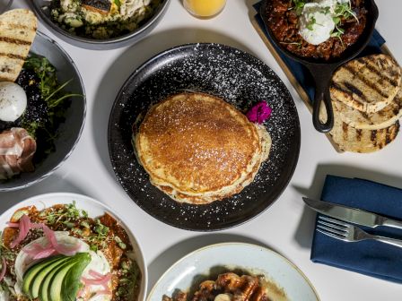 The image shows a variety of dishes including pancakes, salad, grilled bread, and a skillet dish on a table setting.