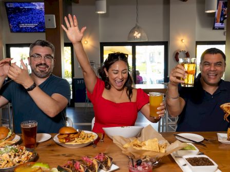 Three people are at a table filled with food and drinks, appearing to be having fun and celebrating, with one person raising arms joyfully.