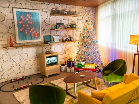 A retro living room with a decorated Christmas tree, vintage TV, toy train set, and colorful furniture, creating a nostalgic holiday atmosphere.