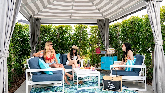 Three people are relaxing under a gazebo with a striped canopy, surrounded by greenery and seating, enjoying a sunny day.