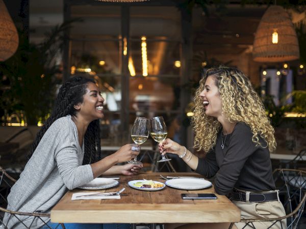 Two women at a restaurant, laughing and toasting with glasses of white wine, enjoying a meal together with plates of food on the table.