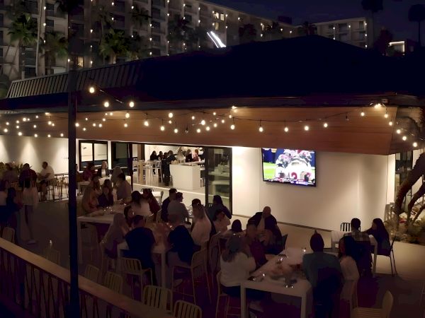 An outdoor restaurant scene at night with diners under string lights, a TV screen showing a sports game, and a modern building background.
