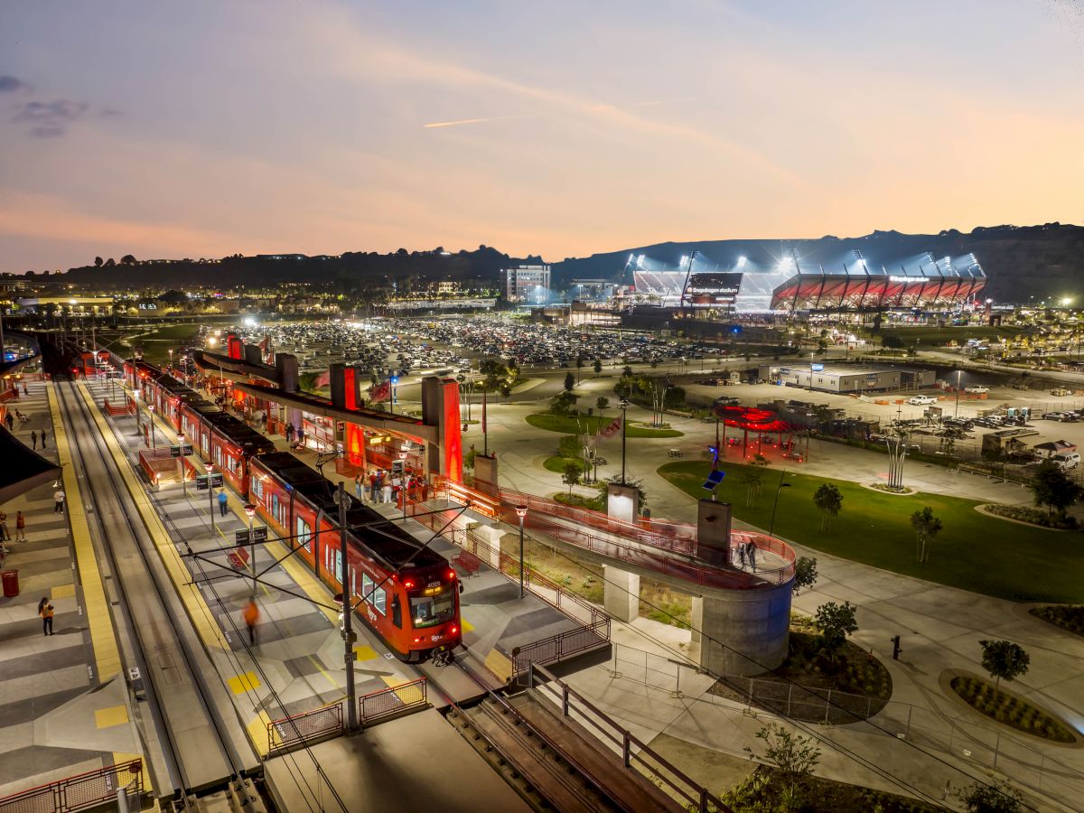 The image shows a transit station with red trains, a parking area, and a stadium in the background against a sunset sky.