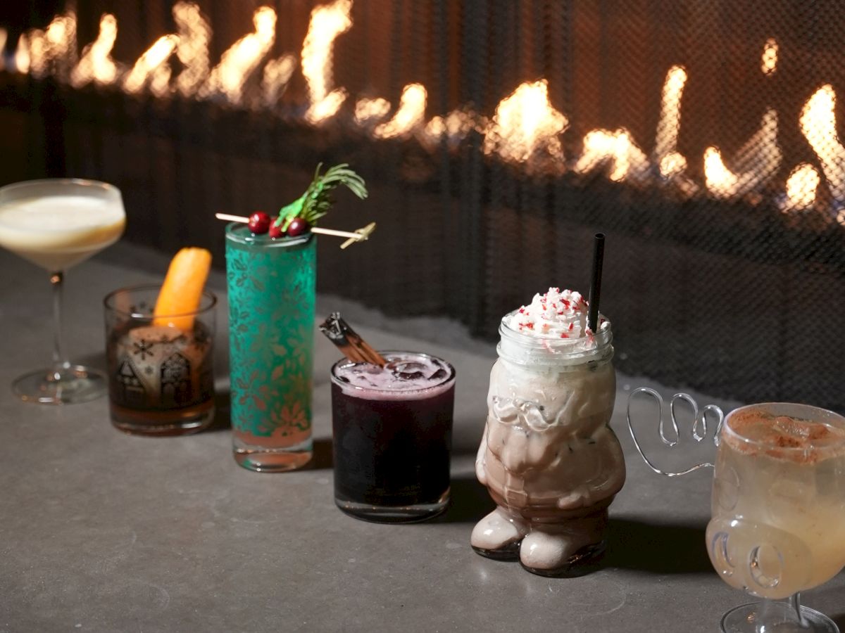 A row of colorful cocktails on a bar counter, with a blazing fireplace in the background and a variety of glasses, including a mint-green drink, a creamy frosted float, and citrus-toned sips, all lined up.
